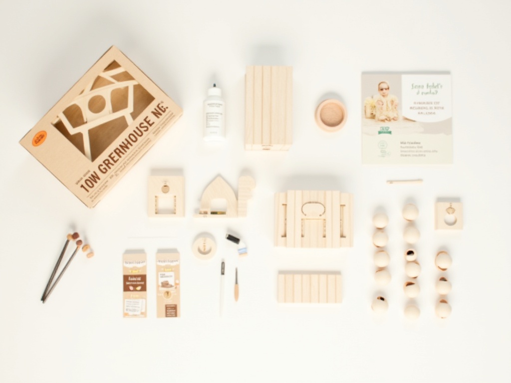 Miniature greenhouse kit parts laid out flat before assembly, including laser-cut wood pieces, acrylic panels, and accessories
